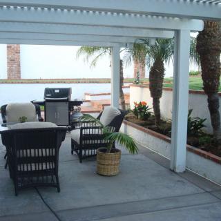 A cozy outdoor patio with a covered pergola, featuring seating and a grill, surrounded by palm trees.