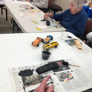 Seniors engaging in a creative painting session, working on toy vehicles at a communal table.