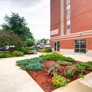A well-landscaped garden area next to a brick building with pathways and greenery.