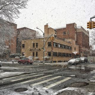 A snowy urban scene depicting a city street with traffic lights and buildings in the background.