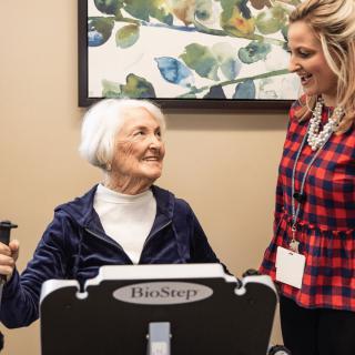 An elderly woman exercises on a machine while interacting with a trainer indoors.