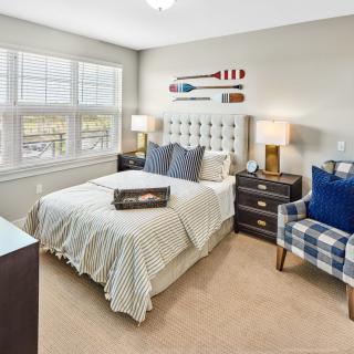 A modern, cozy bedroom featuring a neatly made bed with patterned bedding, a tufted headboard, and bedside tables with lamps. A comfortable chair with blue accents is positioned next to the window, allowing natural light to brighten the room.