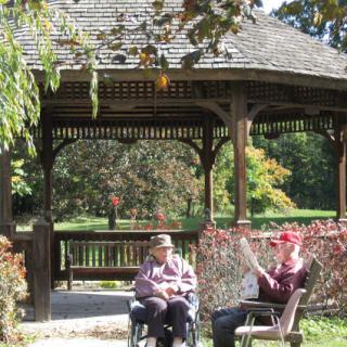 Two elderly individuals enjoying a sunny day in a park under a gazebo.