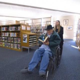 A person in a wheelchair is being pushed by another individual in a library, reading a book.