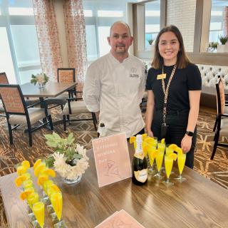 A chef and a staff member stand by a table set with champagne glasses, ready for an event.