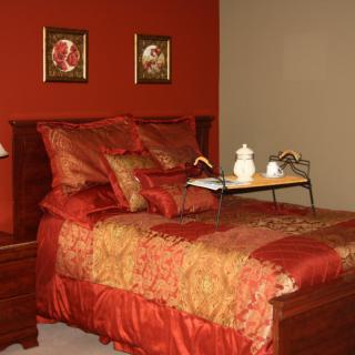 A warmly decorated bedroom with red and gold bedding, featuring artwork on the wall and a bedside table with a lamp.