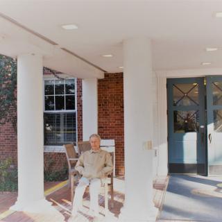A man sitting on a rocking chair on the porch of a brick building with columns.