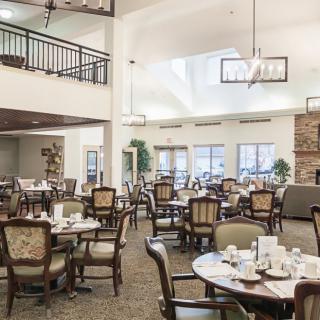 A spacious dining room with elegantly set tables and chairs, illuminated by chandeliers and natural light.