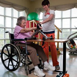 A caregiver assists an elderly woman in a wheelchair with physical therapy exercises.