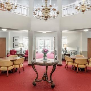 A spacious lobby area with elegant chandeliers, upholstered chairs, and a central table adorned with flowers on a red carpeted floor.
