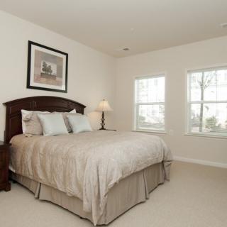 A cozy bedroom featuring a large bed with neutral bedding, flanked by a bedside lamp and artwork on the wall. Natural light streams in through two windows.