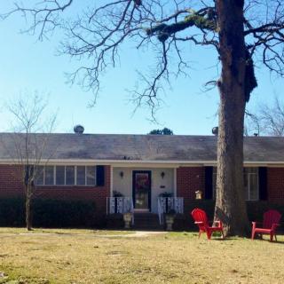 A single-story brick house with a large tree in the front yard and red chairs on the lawn.