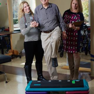 An elderly man participates in a balance exercise with the assistance of healthcare professionals.