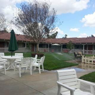 A peaceful patio area with white furniture overlooking a well-maintained lawn and trees under a partially cloudy sky.