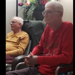 Two elderly men are sitting indoors, appearing relaxed in their chairs.