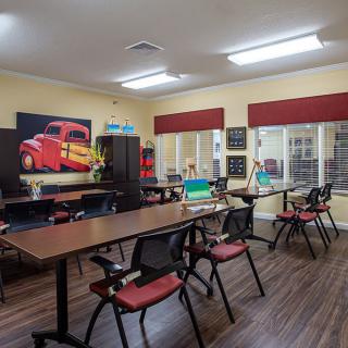 A classroom setting with tables and chairs, featuring artwork on the wall and large windows.