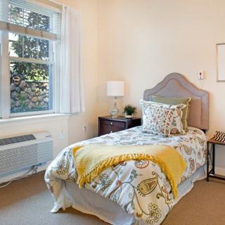 A neatly arranged bedroom with a single bed, decorative pillows, and a nightstand with a lamp near the window.