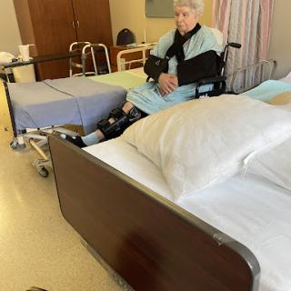 An elderly patient seated in a wheelchair in a hospital room, surrounded by medical equipment and furnishings.