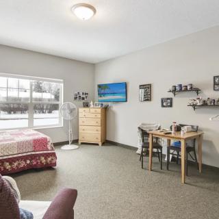 A cozy bedroom interior with a bed, dresser, table, and chairs, featuring wall decorations and a large window.