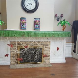 A fireplace adorned with tiki-themed decorations, featuring a clock, green fringe, and spider webs.
