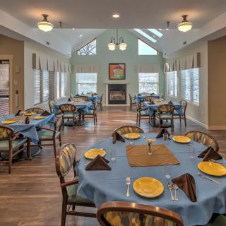 A cozy dining room with tables set for a meal, featuring blue tablecloths and ample natural light.
