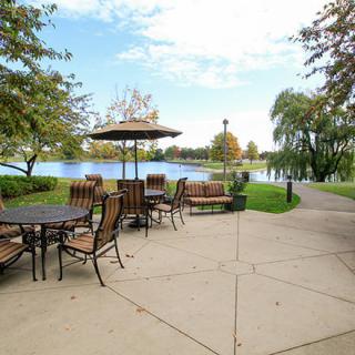 A serene outdoor patio overlooking a lake with tables and chairs surrounded by trees and a winding pathway.