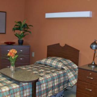 A cozy bedroom with a single bed, orange walls, and simple furnishings including a nightstand with a lamp and a vase of flowers.