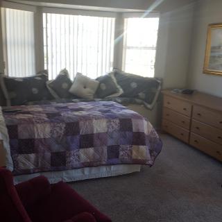 A cozy bedroom featuring a quilt-covered bed with multiple pillows, flanked by a window letting in natural sunlight. A dresser and soft carpet complete the decor.