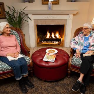 Two seniors sitting comfortably by a fireplace, engaged in conversation.