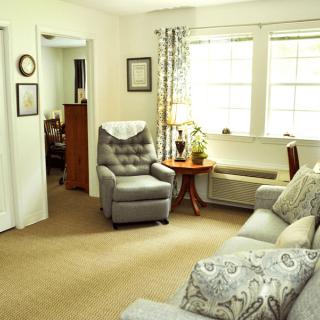 A cozy living room with a comfortable armchair and sofa arranged around a small table, well-lit by natural light through large windows.