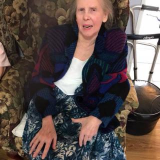 An elderly woman sitting comfortably in an armchair indoors, wearing casual clothing.