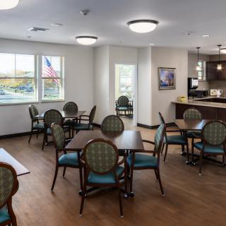 A cozy dining area with several tables and chairs, adjacent to a kitchen space, featuring large windows and overhead lighting.