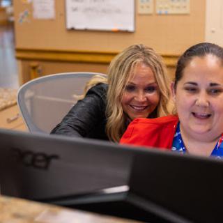 Two women are working together at a computer in an office setting, sharing a moment of joy.