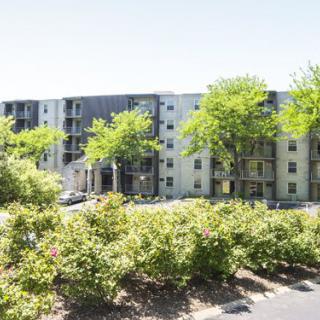 A modern apartment building surrounded by trees and a parking lot on a sunny day.