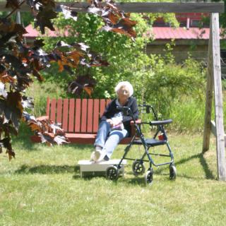 An elderly person sitting on a bench outdoors with a walker nearby.