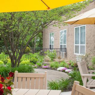 A tranquil garden patio with a table, chairs, and yellow umbrellas surrounded by lush greenery.