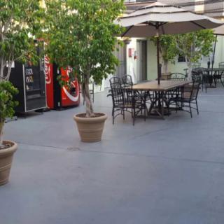 An outdoor patio area with tables, chairs, and umbrellas, complemented by potted plants and a nearby vending machine.
