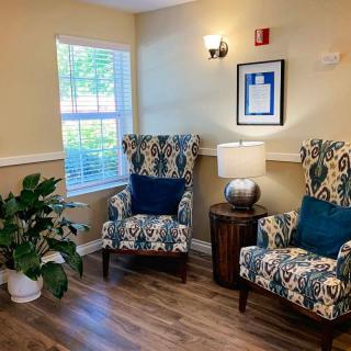 A cozy living room area featuring two patterned chairs, an indoor plant, a side table with a lamp, and a framed artwork on the wall.