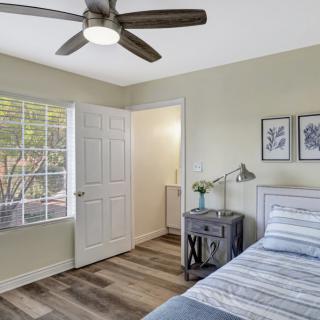 A cozy bedroom with a bed, side table, and ceiling fan, featuring natural light from a large window.