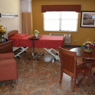 A well-furnished hospital room with a bed, table, chairs, and decorative flowers.