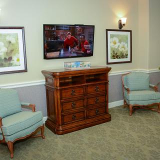 A cozy living room with armchairs and a television mounted above a wooden cabinet, flanked by framed paintings.