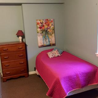 A cozy bedroom with a single bed adorned with a pink bedspread, next to a wooden dresser and floral artwork on the wall.