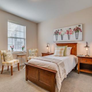 A cozy bedroom with a wooden bed, two chairs by the window, and floral artwork on the wall.