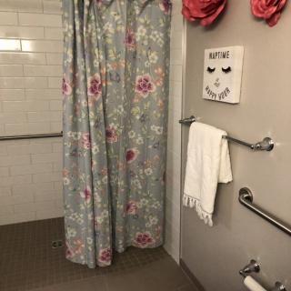 A bathroom with a floral shower curtain and handrails, featuring a towel rack and decorative wall art.