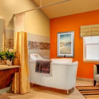 A modern bathroom with orange walls, featuring a standalone bathtub, artwork on the wall, a window with blinds, and decorative plants.