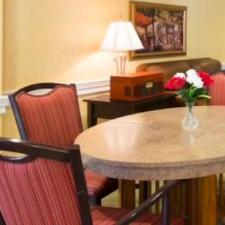 A cozy dining area featuring a round wooden table with red upholstered chairs, a vase of roses, a lamp, and a painting on the wall.