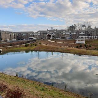 A serene view of a pond reflecting the cloudy sky, surrounded by buildings and greenery.