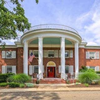 A grand mansion with large white columns and a red door, set amidst lush greenery.