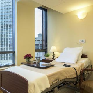 A hospital room featuring a neatly made bed with a city view through large windows.