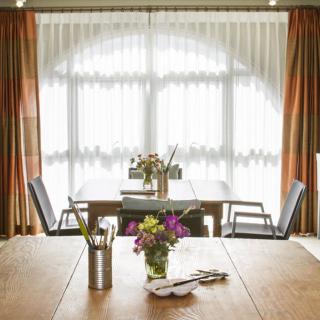 A sunlit dining area with a wooden table, chairs, and vases of flowers by the window.